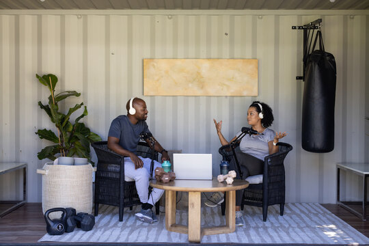Diverse cohosts recording podcast in container studio wearing athletic wear with mics and laptop
