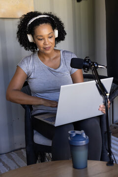 African American woman recording on laptop in home studio, wearing white headphones and using mic
