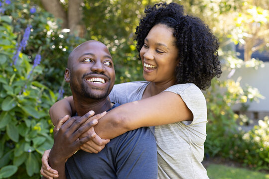 African American couple sitting in garden, woman hugging man's shoulders, purple flowers nearby