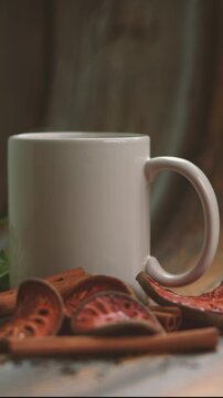 A steaming white ceramic mug of hot herbal tea sits on a dark, rustic wooden surface, surrounded by fragrant cinnamon sticks and dried bael fruit slices