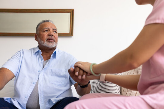 Senior African American man sitting on sofa while Asian caregiver in pink scrubs holding hand