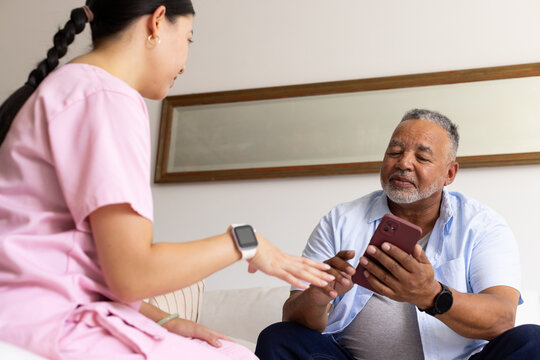 Senior African American man using red phone on sofa Asian woman caregiver in pink-scrubs guiding