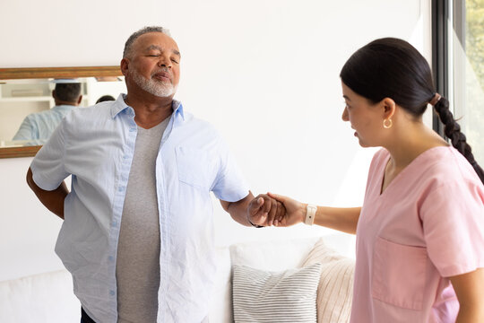 Senior African American man receiving support from Asian caregiver in pink scrubs in living room