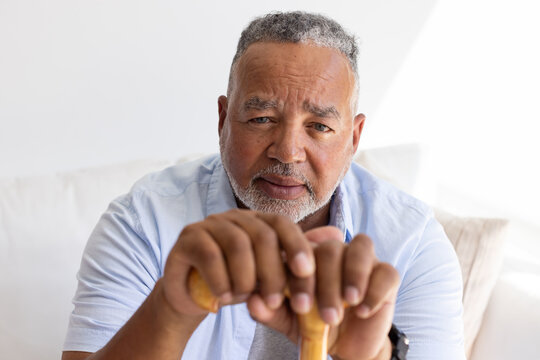 Senior African American man sitting on sofa holding wooden cane in living room wearing blue shirt
