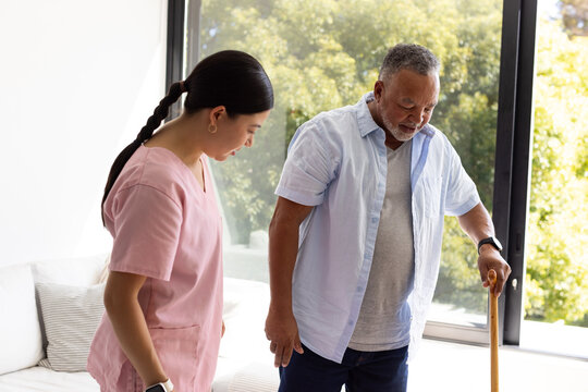 Diverse senior man using wooden cane while caregiver in pink scrubs assisting near window and sofa