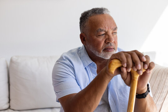 Senior African American man sitting on pale sofa wearing casual shirt and wristwatch holding cane