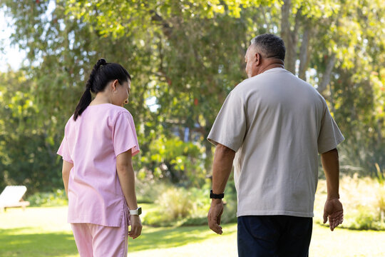 Diverse pair walking on lawn Asian nurse in pink scrubs showing smartwatch, man with watch, bench