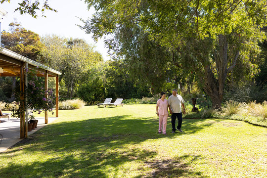 Diverse couple walking side-by-side on sunlit backyard lawn, woman wearing pink scrubs near pergola