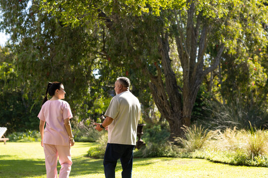 African American senior man, Asian woman caregiver in pink scrubs walking across garden near urn