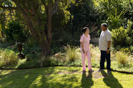 African American man and Asian caregiver in pink scrubs talking on lawn in garden with watches