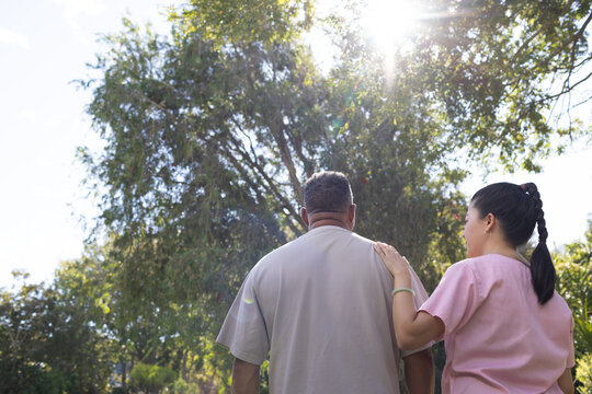 African American man with Asian nurse placing hand on shoulder beside park tree in pink scrubs