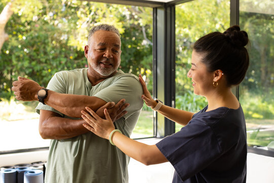 Diverse therapist guiding mature male client stretching shoulder in living room with rolled mats