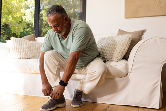 Senior African American man tying shoe laces in living room on cream sofa with smartwatch