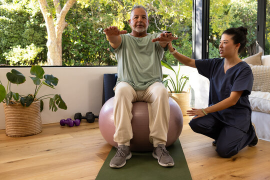 Senior African American man balancing on exercise ball at home, Asian therapist in scrubs guiding