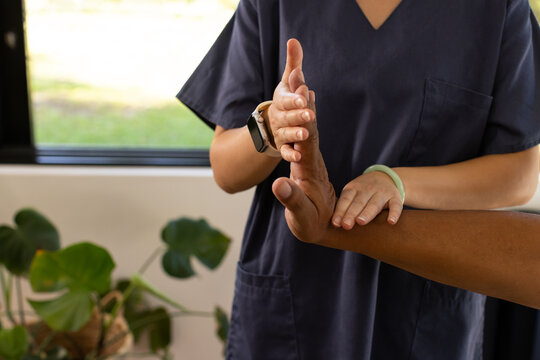 Asian female clinician supporting male patient's wrist in navy scrubs beside window with smartwatch