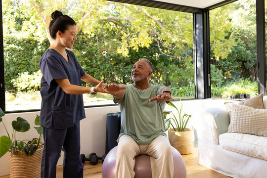 Asian therapist in navy scrubs helping senior African American man on purple exercise ball at home