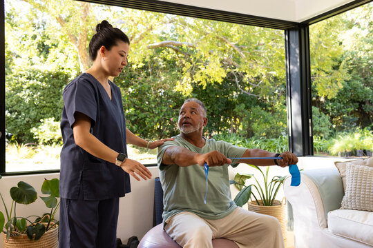 African American man, caregiver in navy scrubs guiding row on ball using blue band at home