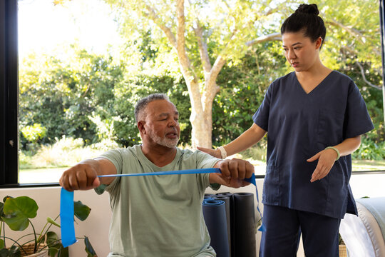 Diverse therapist in scrubs guiding patient doing chest press with blue band by window, copy space