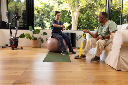 Asian therapist adjusting watch as African American male patient using yellow band in living room