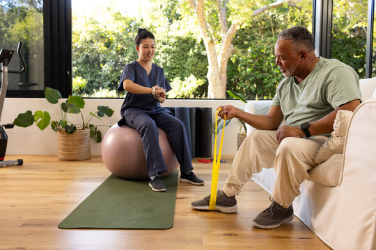 Diverse therapist in navy scrubs guiding client in living room using yellow resistance band