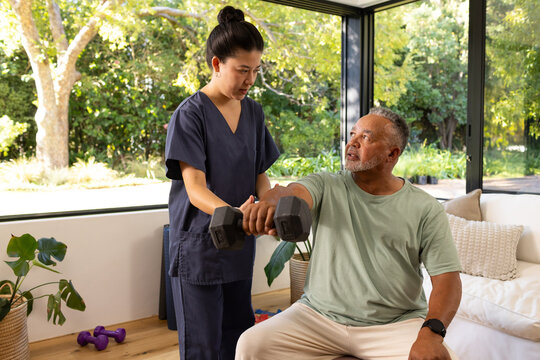 Senior African American man lifting hex dumbbell while Asian nurse in scrubs helping in living room