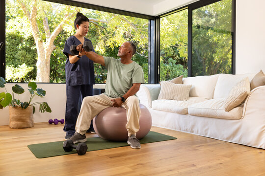 Senior African man and Chinese woman in navy-scrubs guiding dumbbell curl on stability ball in home