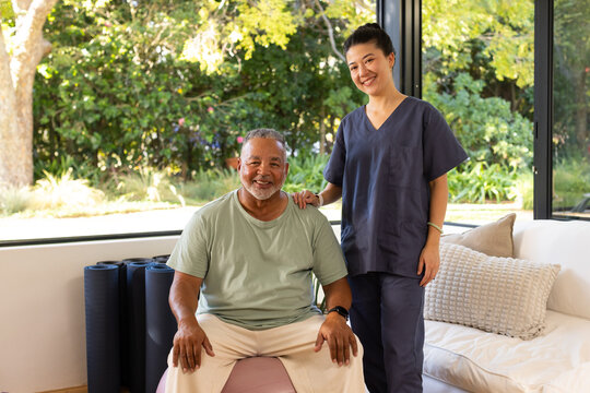 African-American senior sitting on pink ball at home Asian nurse in scrubs supporting shoulder