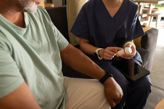 Mature African American man and Asian woman scrub-clad sitting on couch holding pill bottle, tablet