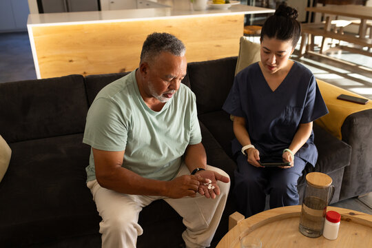 Diverse female caregiver in navy scrubs checking tablet on sofa while male patient holding pills