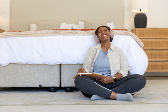 Woman sitting cross-legged on bedroom floor laughing and writing in notebook with headphones