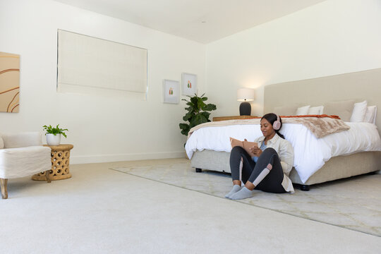 Woman sitting in bedroom by large bed writing in notebook in leggings and headphones, copy space