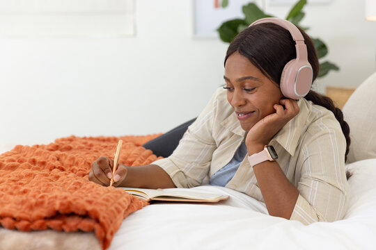 African American woman lying on bed writing in open notebook with pink headphones, copy space