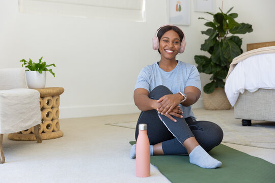Adult African woman sitting on green yoga mat in bedroom wearing pink headset, holding water bottle