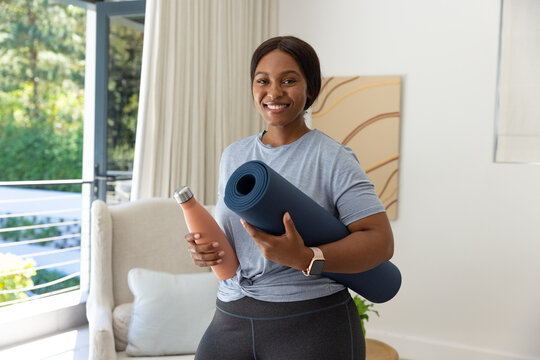 African woman standing at home holding blue yoga mat and coral bottle, wearing gray tee, smartwatch