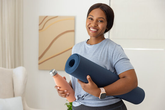 African woman standing in living room holding yoga mat and peach bottle, wearing knotted tee