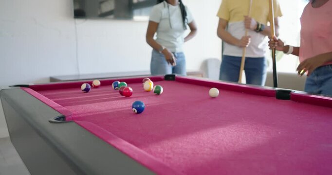 Diverse friends gathering at rec room pink-felt table after break, woman in pink top executing shot