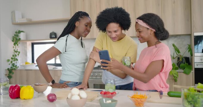 African American friends cooking at kitchen island, friend in pink showing smartphone, sharing clip