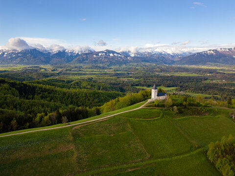 Aerial view of the Church of St. Primus and Felician on a green hilltop ridge with snow-capped mountains in the background Jamnik, Kranj, Slovenia.