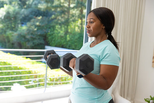 African woman lifting black hex dumbbells wearing light aqua athletic tee near sliding glass door