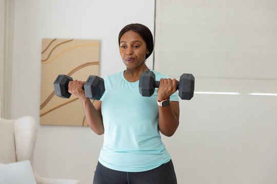 Woman in athletic top and leggings holding hex dumbbells and performing bicep curls at home