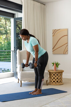 Woman hinging on blue yoga mat in living area wearing turquoise top holding hex dumbbells