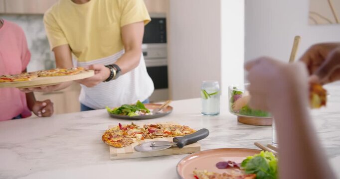 African American friends sharing pizza at kitchen counter, man carrying board, woman cutting slices