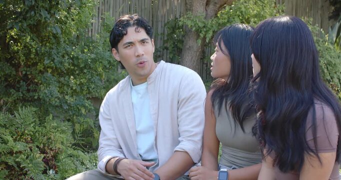 Three friends sitting on seat in garden man making point gesturing women listening watch visible