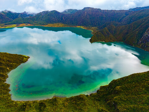 Aerial view of Lagoa do Fogo crater lake with turquoise water reflecting white clouds surrounded by lush green volcanic mountains in Agua de Alto, Azores, Portugal.