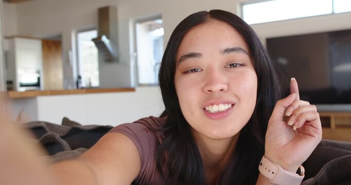 Asian woman reaching camera, adjusting it, leaning in living room in mauve top, speaking to camera
