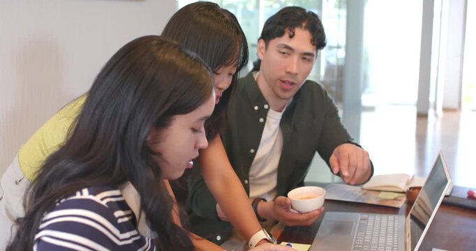 Diverse coworkers working on task at table, man pointing at laptop holding bowl, women taking notes