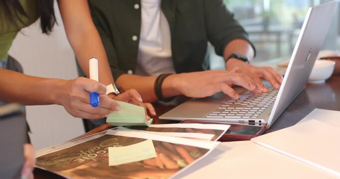 Diverse team reviewing prints at table, writing sticky notes, pointing at laptop, typing for review