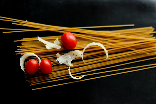  Raw spaghetti with cherry tomatoes and parmesan cheese on a black background. Minimalist food composition with dramatic lighting, highlighting textures and natural colors. 