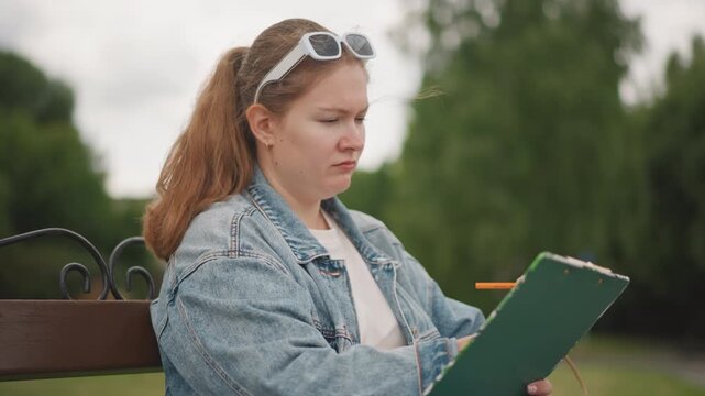Caucasian art student sketching on bench focused on sketchbook and pencil, denim jacket, sunglasses on head, backpack at side, overcast sky and lush greenery creating quiet reflective mood