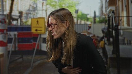 Fototapeta premium Woman clutching stomach with hand and wincing on street near barricade; distress pain urgency emergency.